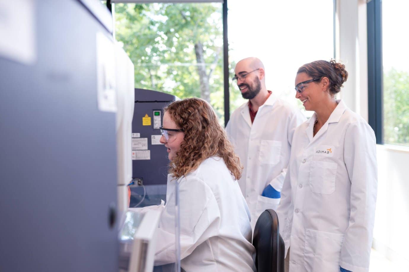 Three scientists in lab coats working together at an antibody discovery laboratory workstation.