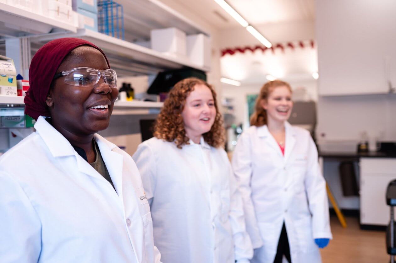 Three scientists in lab coats talking in a laboratory setting.